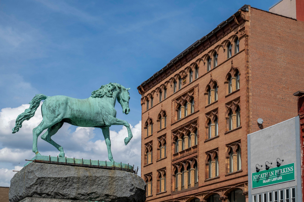 A bronze horse statue stands on a stone pedestal in front of a historic brick building with arched windows. The sky is blue with scattered clouds.